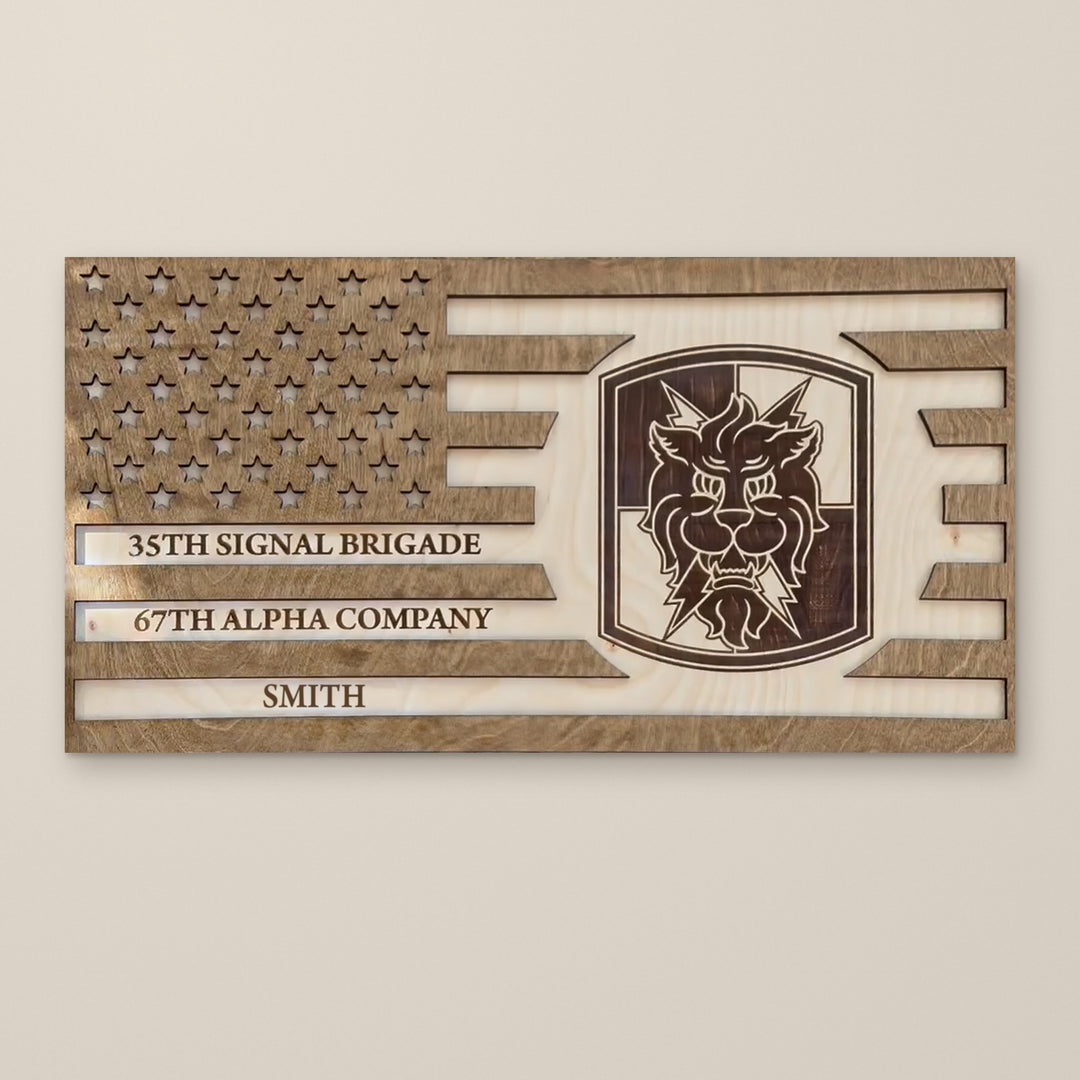 Custom American flag sign in wood with three engraved lines, cut-out stars and a unit insignia, shown on a wall (sample name SMITH)